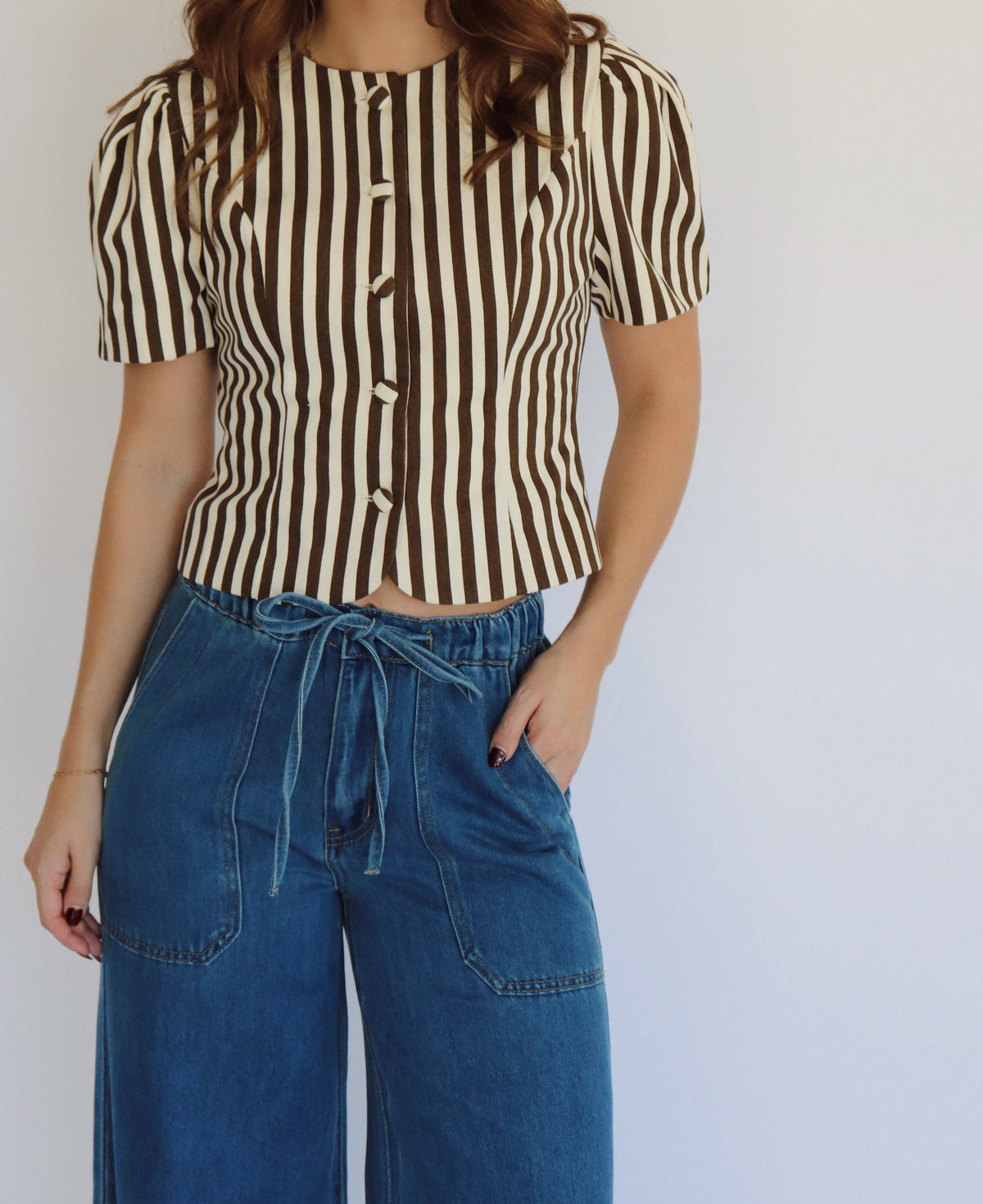 A woman wearing a brown and beige vertically striped short-sleeve button-up top with blue wide-leg jeans, standing against a plain light gray wall.