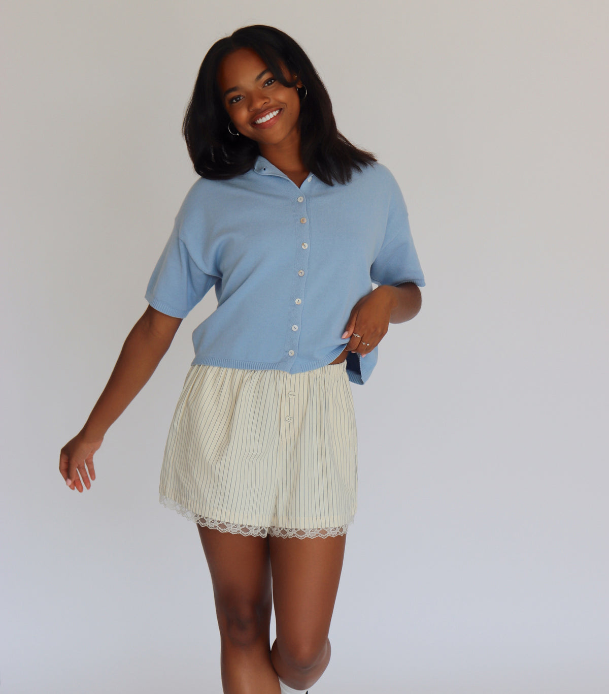 Woman wearing a blue short sleeve cardigan and cream shorts against a white background