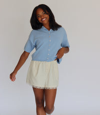 Woman wearing a blue short sleeve cardigan and cream shorts against a white background