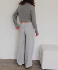 A person shown from the back wearing a light gray long-sleeve cropped sweater and wide-leg sweatpants, standing indoors against a white wall with a light-colored bench and wood floor visible.