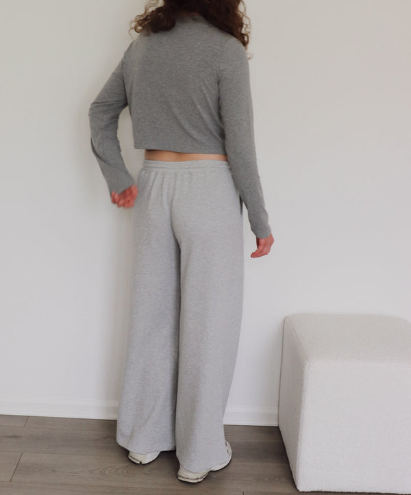 A person shown from the back wearing a light gray long-sleeve cropped sweater and wide-leg sweatpants, standing indoors against a white wall with a light-colored bench and wood floor visible.