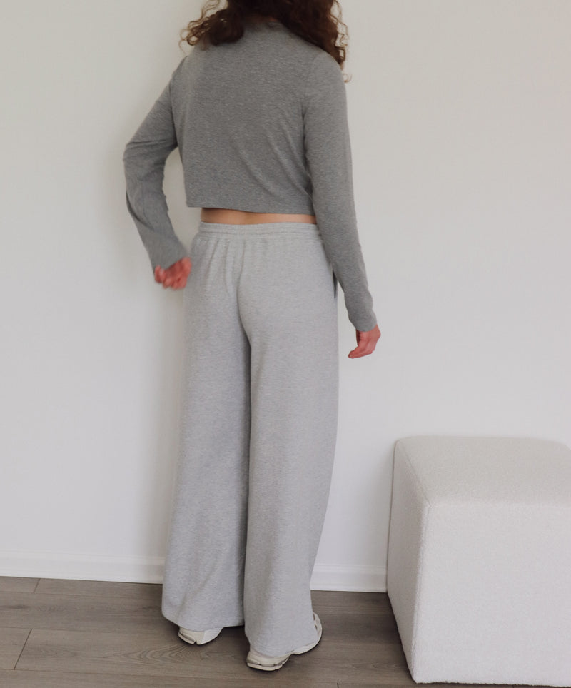 A person shown from the back wearing a light gray long-sleeve cropped sweater and wide-leg sweatpants, standing indoors against a white wall with a light-colored bench and wood floor visible.