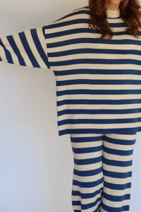 A woman wearing a matching two-piece loungewear set with blue and beige horizontal stripes, featuring a long-sleeve top and wide-leg pants, standing against a plain white background.