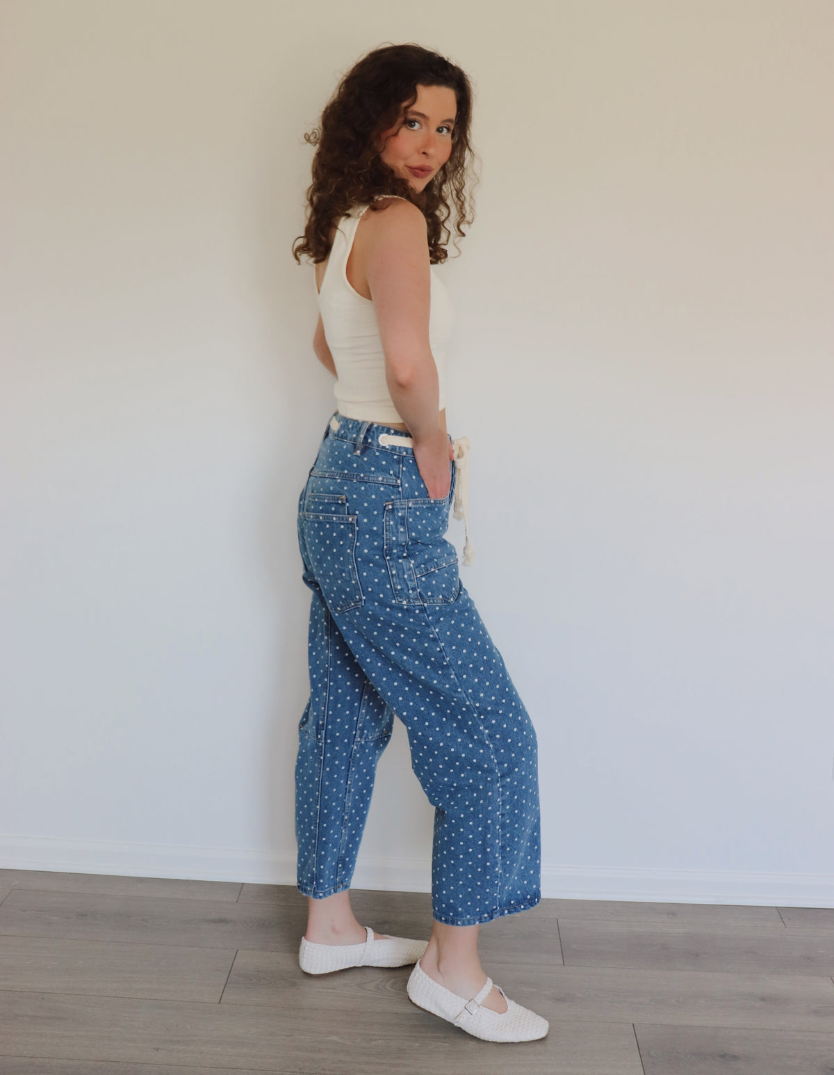 woman wearing white tank top and blue jeans with white polka dots on them against a white wall