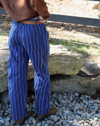 A person viewed from behind wearing blue denim jeans with thin vertical stripes, a brown sweater, and brown boots, standing outdoors on a bed of white gravel near large rocks.