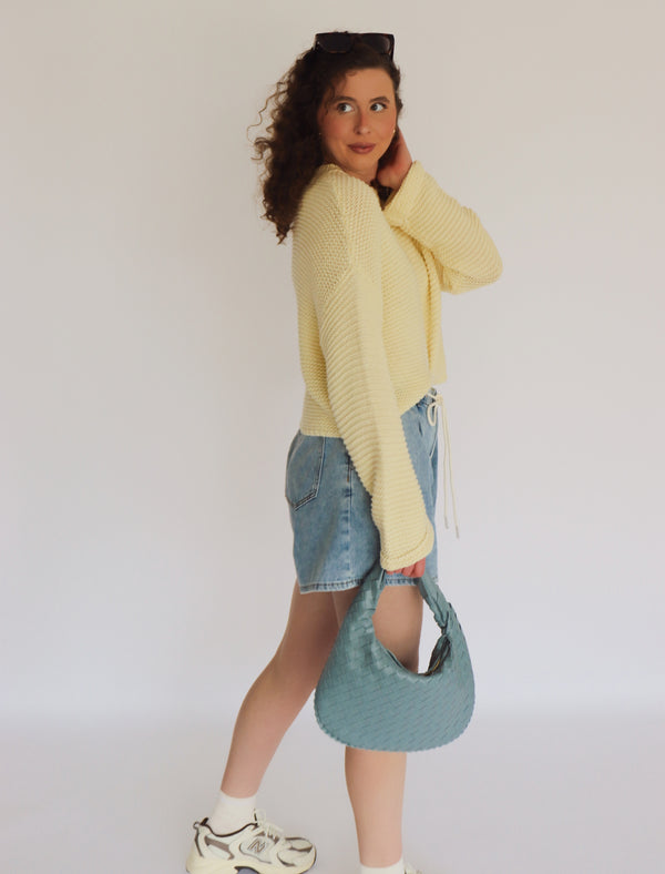 Woman wearing a yellow sweater and denim shorts, holding a blue bag against a white background
