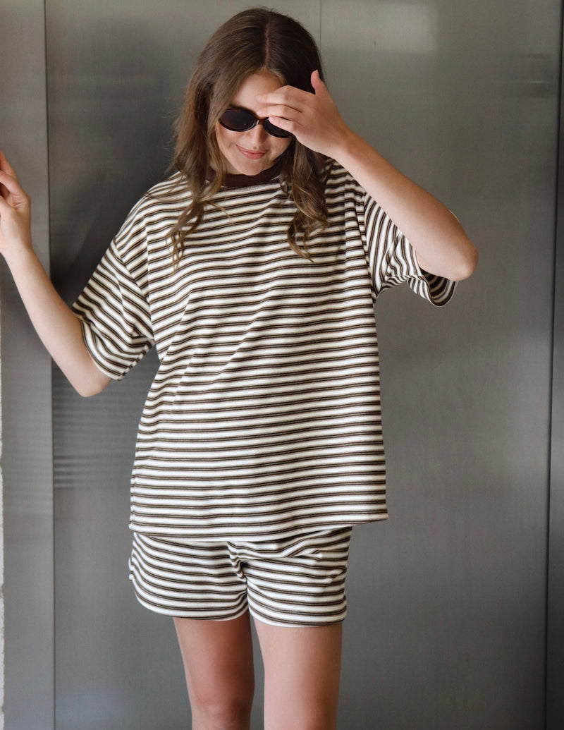 A woman wearing an oversized short-sleeve striped t-shirt and matching striped shorts, posing against a dark gray paneled wall.