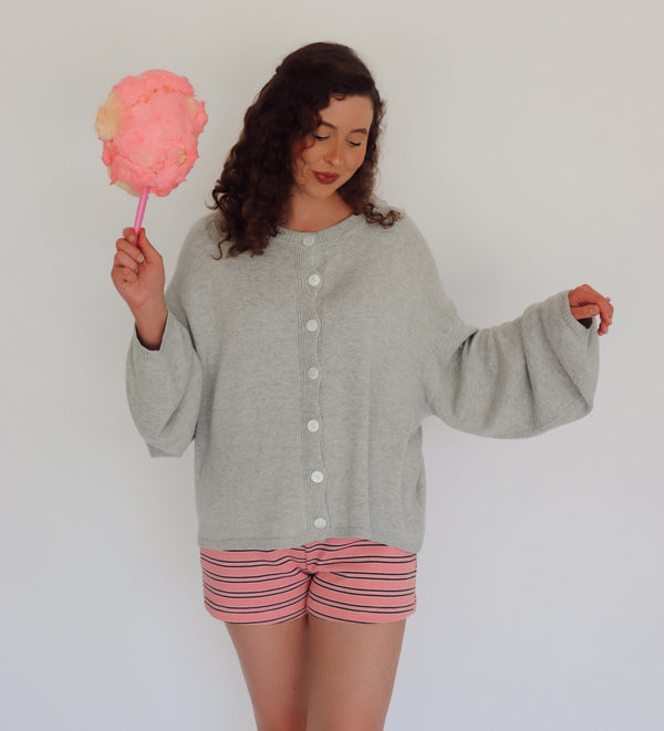 A woman wearing a light gray knit cardigan and pink striped shorts holds a pink cotton candy against a plain white background.