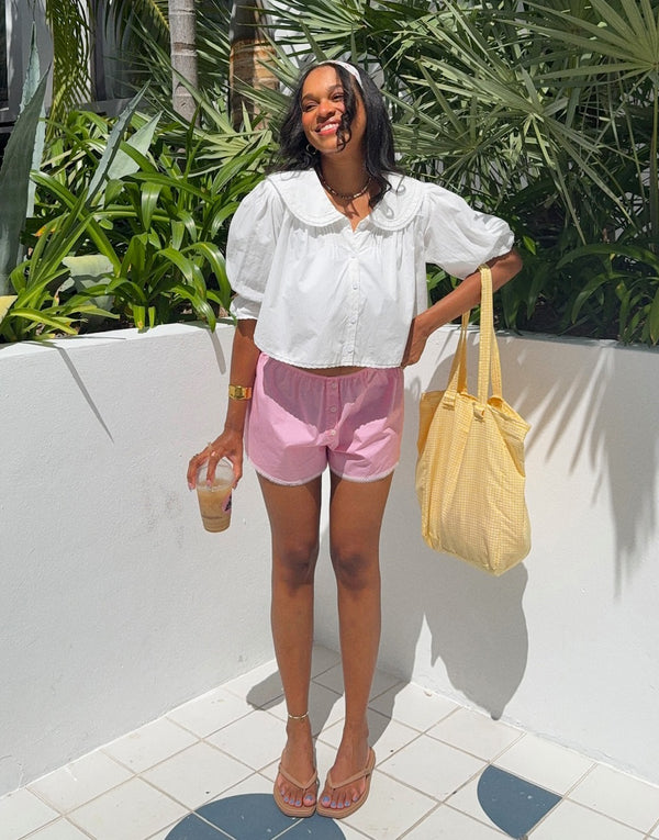 Woman in white blouse and pink shorts holding a yellow bag and drink, standing in front of green plants.