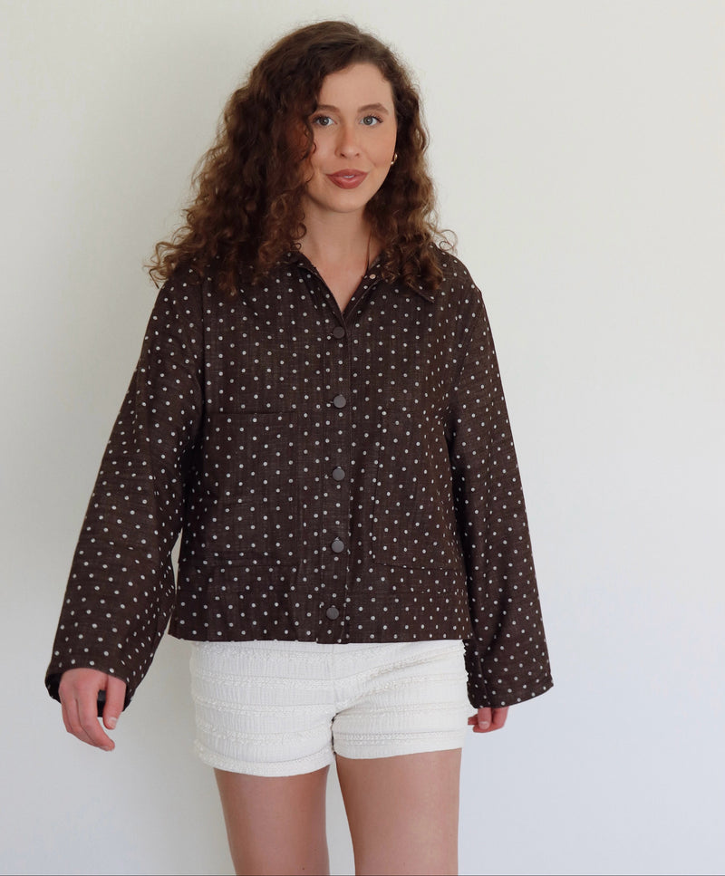 A woman with curly brown hair wearing a dark brown long-sleeve button-up jacker with a white polka-dot pattern and white textured shorts, standing against a plain off-white wall.