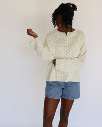 Woman wearing a cream/light gray sweater with small pointelle patterns and denim shorts on a white background