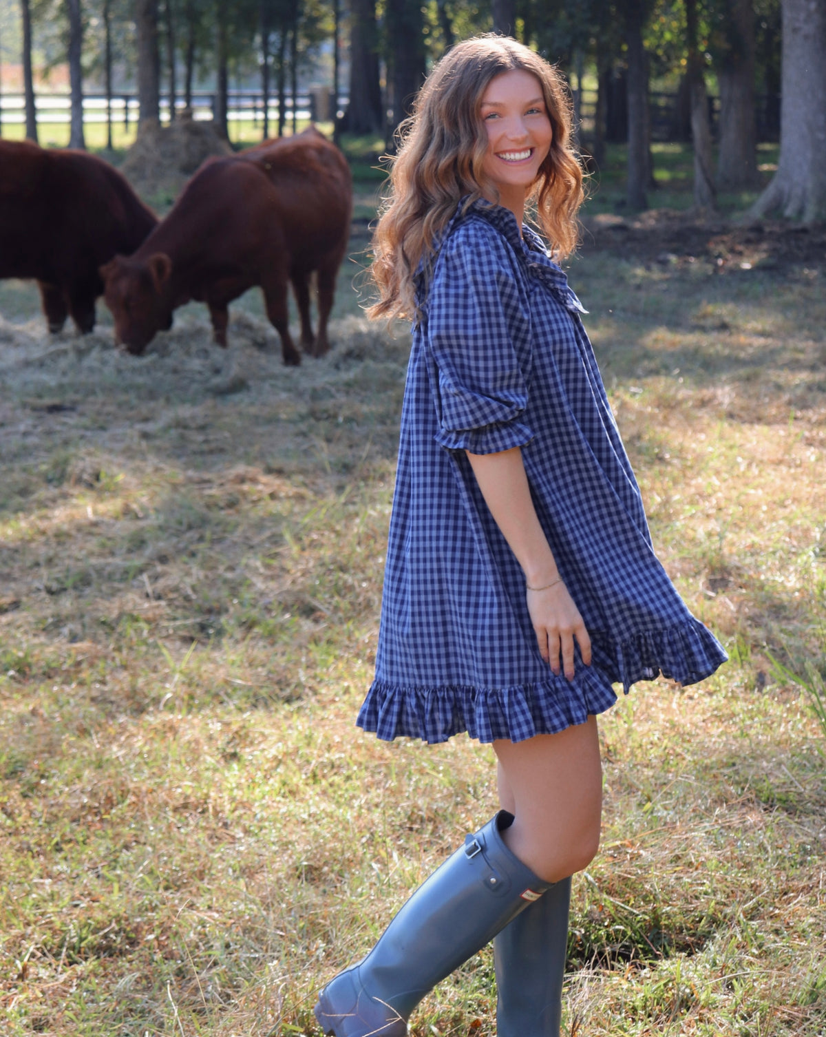 A woman wearing a blue gingham puff-sleeve dress with a ruffled neckline and hem, standing in a grassy field with cows in the background. She is wearing rain boots.