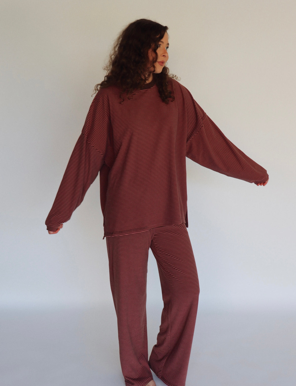 A woman modeling a matching two-piece loungewear set consisting of a long-sleeve top and pants, both featuring thin horizontal stripes in a pink and dark brown colorway.