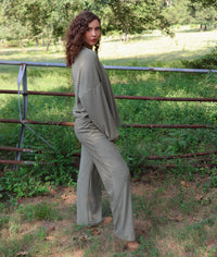 A woman wearing a matching olive green striped lounge set stands outdoors in a wooded area with greenery and a rustic fence behind her.