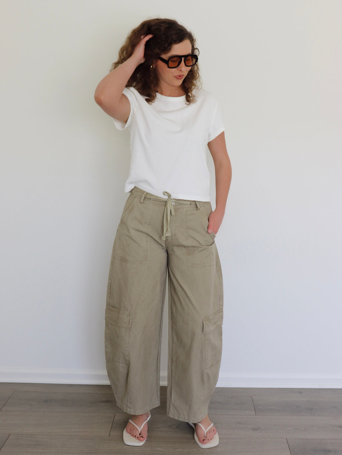 A woman wearing a white t-shirt, wide-leg beige cargo pants, and white flip-flops stands against a plain white wall on a light wood floor.