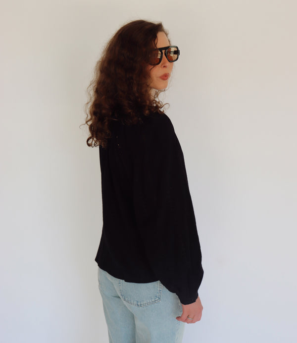 A woman wearing a black long-sleeve blouse with lace and embroidered shoulder details, paired with light blue jeans and black sunglasses, posing against a plain white background.