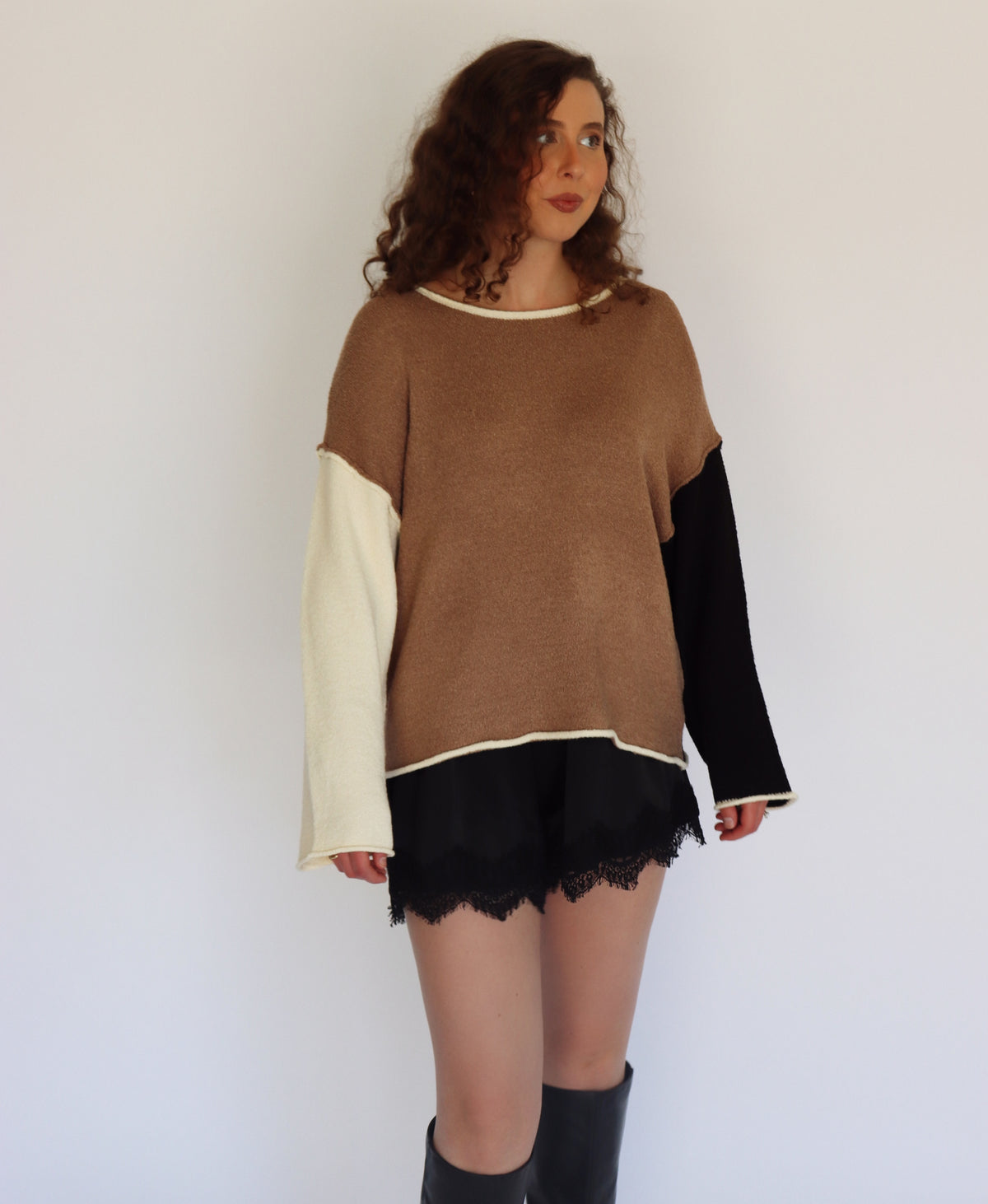 A woman modeling a color-block knit sweater with a brown body, black sleeves, and cream-colored underarms and cuffs, paired with black shorts and black knee-high boots against a plain white background.