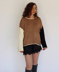 A woman modeling a color-block knit sweater with a brown body, black sleeves, and cream-colored underarms and cuffs, paired with black shorts and black knee-high boots against a plain white background.