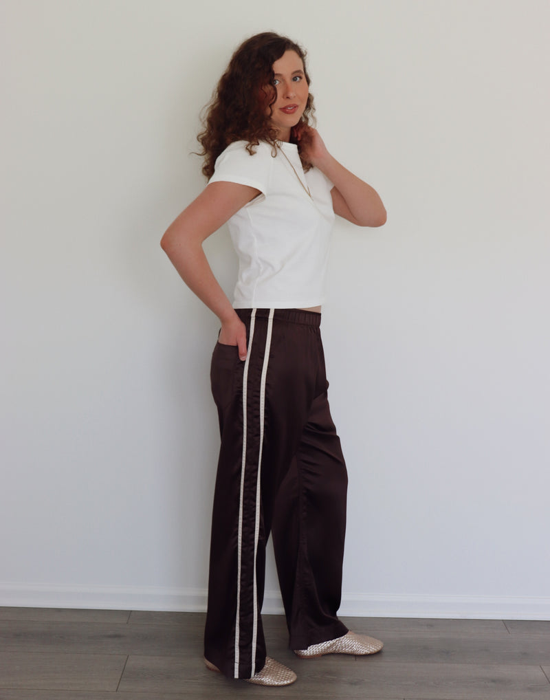 A woman with curly brown hair stands in profile wearing dark brown wide-leg pants with a white stripe down the side, a white cropped t-shirt, and beige flats, set against a plain white wall and light wood floor