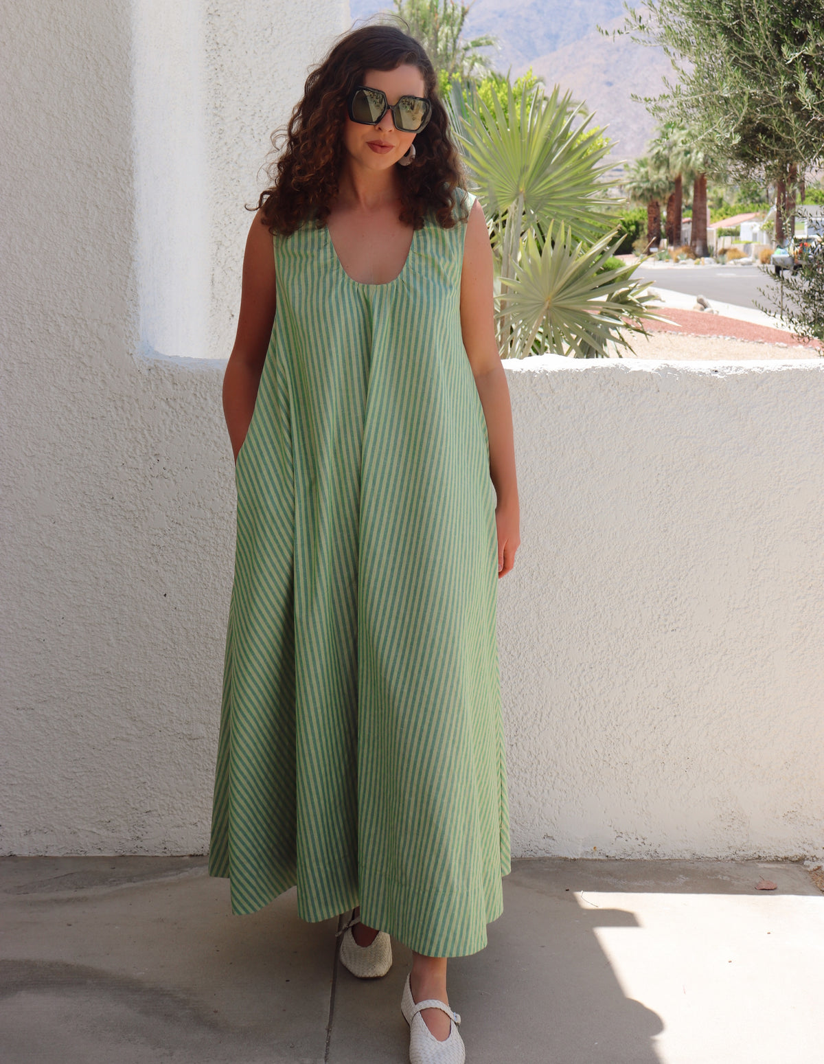 Woman wearing a green striped dress standing against a white wall with plants in the background.