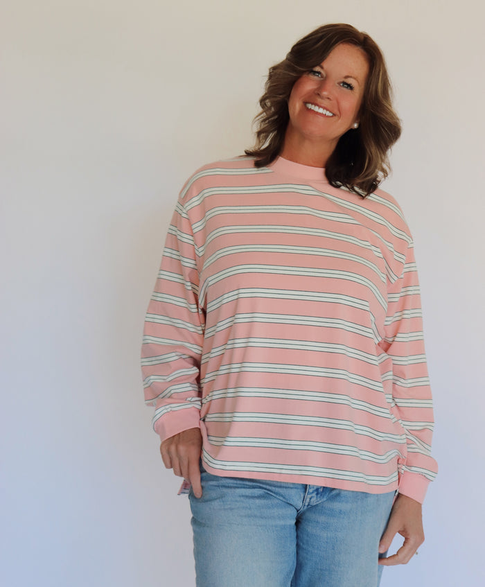 A woman wearing a pink long-sleeve crewneck shirt with thin white horizontal stripes, paired with light blue jeans, standing against a plain off-white wall.