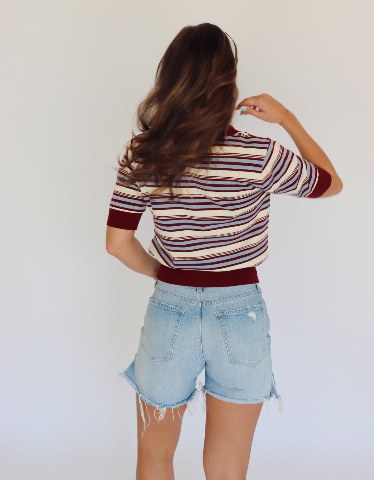 A woman shown from behind wearing a short-sleeve striped knit top with burgundy, light blue and cream horizontal stripes, paired with light-wash denim shorts, against a plain white background.