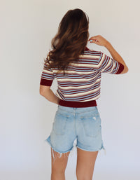 A woman shown from behind wearing a short-sleeve striped knit top with burgundy, light blue and cream horizontal stripes, paired with light-wash denim shorts, against a plain white background.
