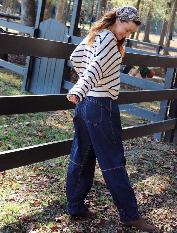 A woman wearing a cream and navy striped long-sleeve shirt and dark blue denim work pants, standing outdoors in front of a wooden fence.
