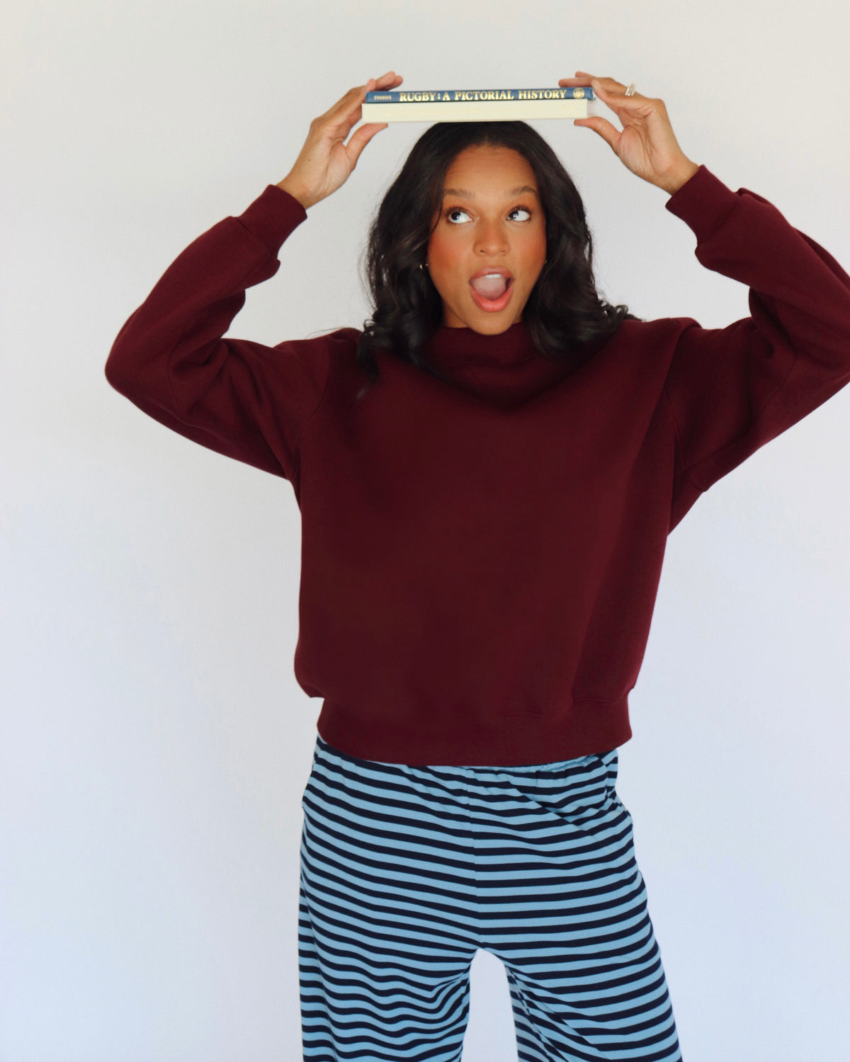 A woman posing in a burgundy mock neck and blue-and-navy striped pants, holding a book above her head against a plain white background.