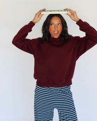 A woman posing in a burgundy mock neck and blue-and-navy striped pants, holding a book above her head against a plain white background.