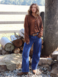 A woman stands outdoors next to a tree and a wooden fence, wearing a brown button-up cardigan and blue striped pants.