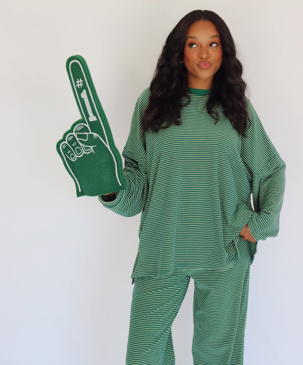 A woman wearing a green and yellow  striped long-sleeve top and matching pants, holding a green foam finger with white '#1' text, against a plain white background.