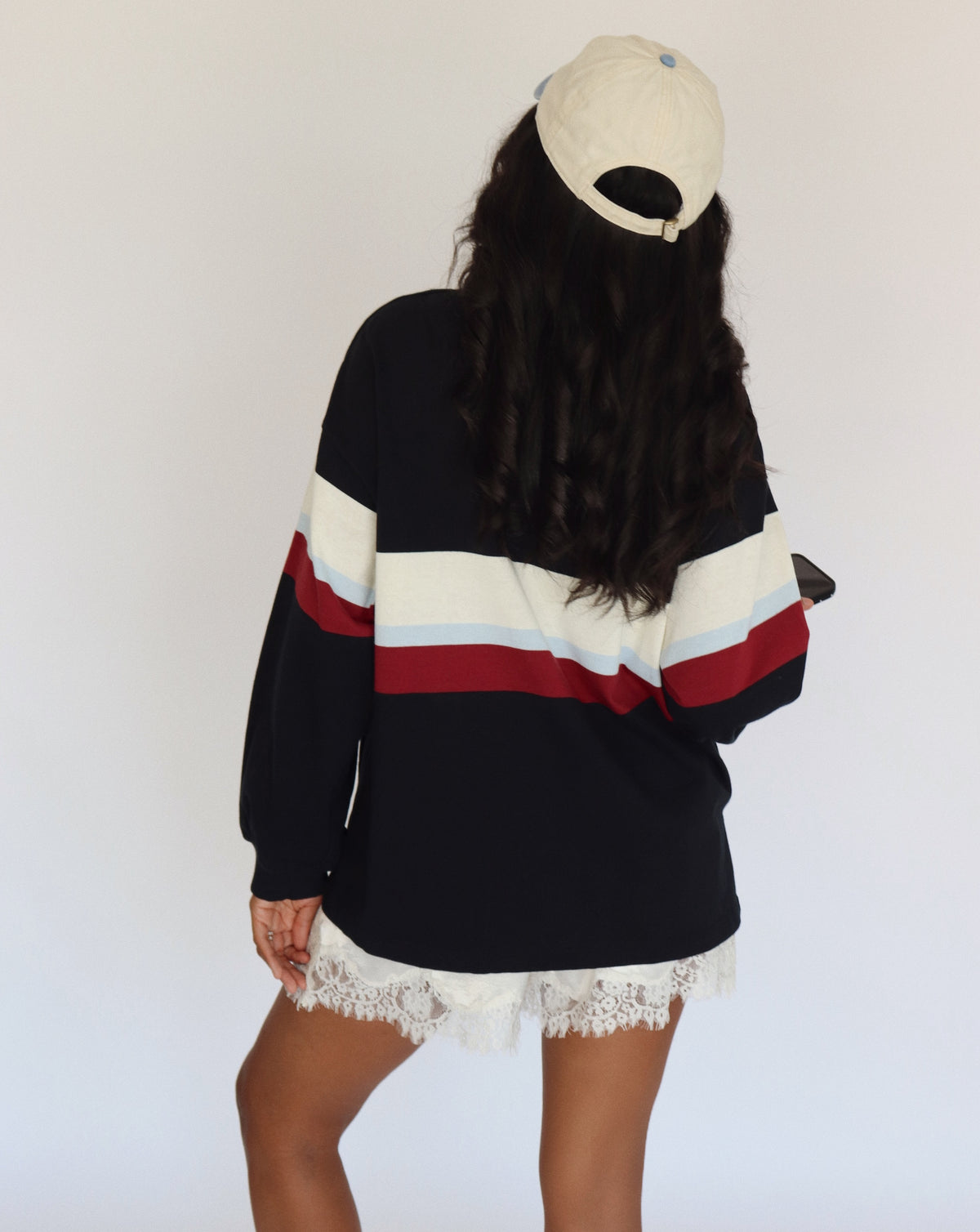 A woman shown from behind wearing a black long-sleeve sweatshirt with wide cream, light blue, and red horizontal stripes across the chest and sleeves, paired with white lace-trim shorts and a cream baseball cap.