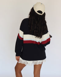 A woman shown from behind wearing a black long-sleeve sweatshirt with wide cream, light blue, and red horizontal stripes across the chest and sleeves, paired with white lace-trim shorts and a cream baseball cap.