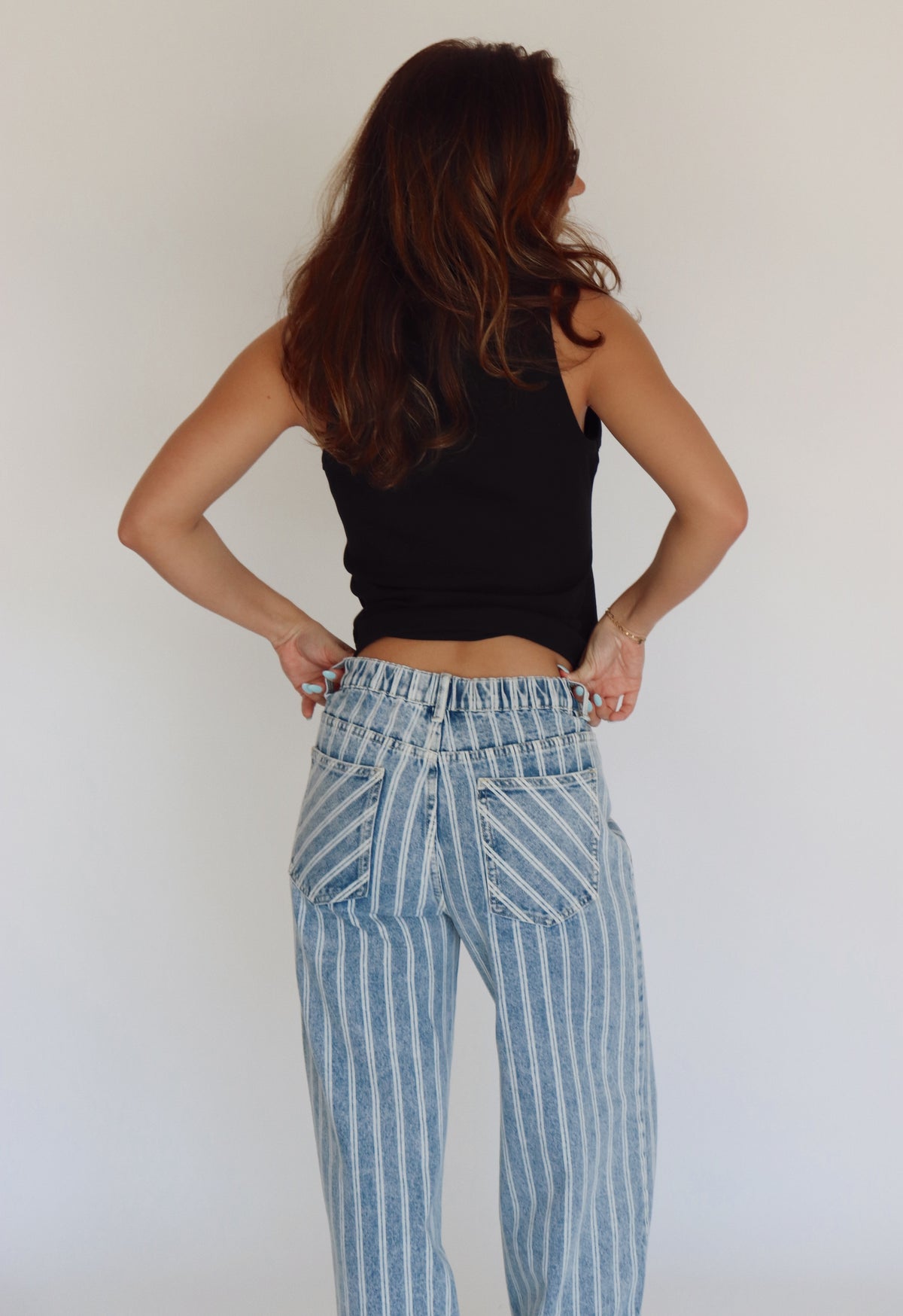 A woman shown from behind wearing light blue and white vertically striped high-waisted jeans and a black tank top, posing with hands on hips against a plain white wall.