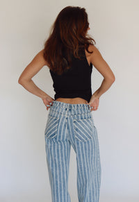 A woman shown from behind wearing light blue and white vertically striped high-waisted jeans and a black tank top, posing with hands on hips against a plain white wall.