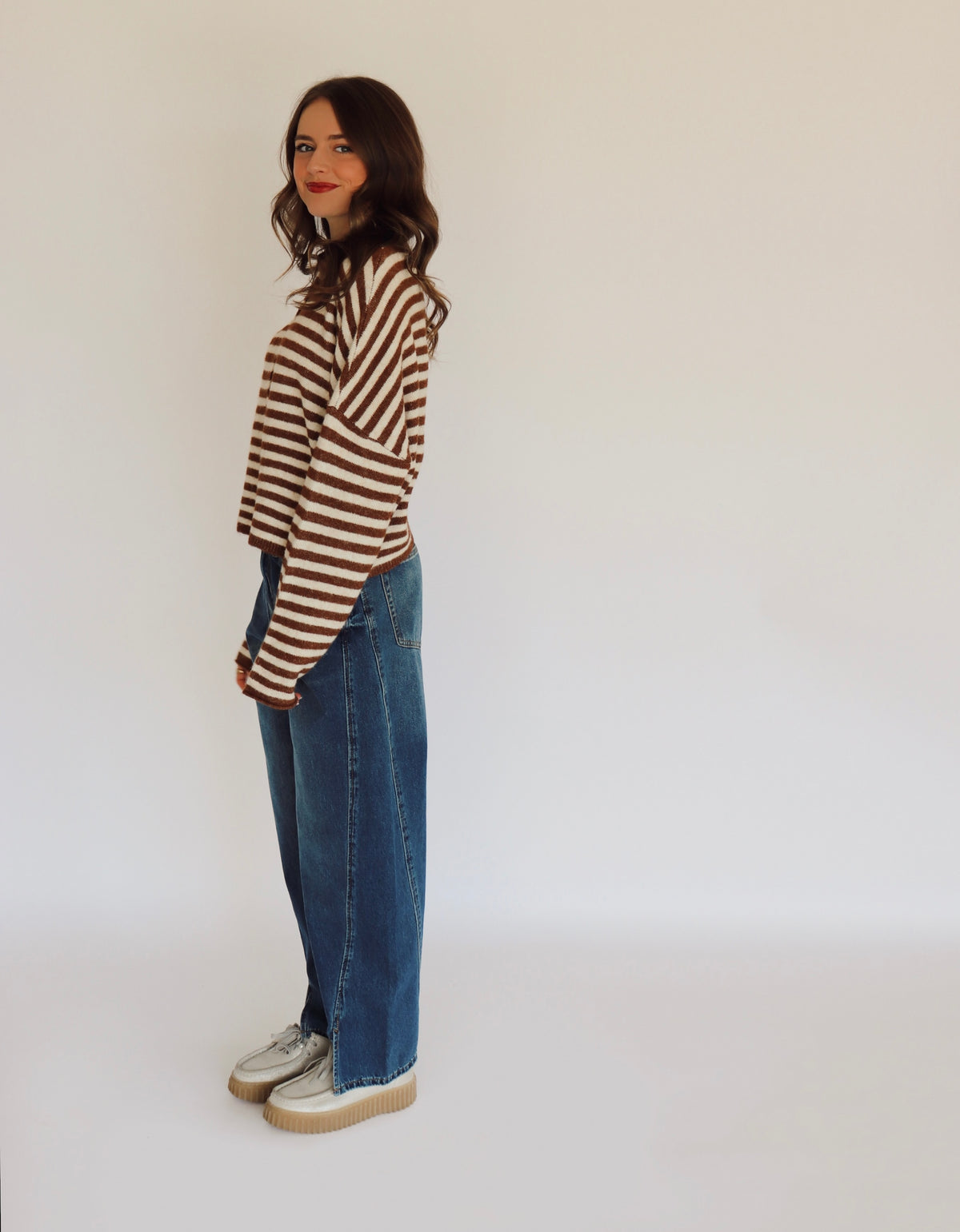 Woman wearing a brown and white striped sweater cardigan and blue jeans in front of  a white background