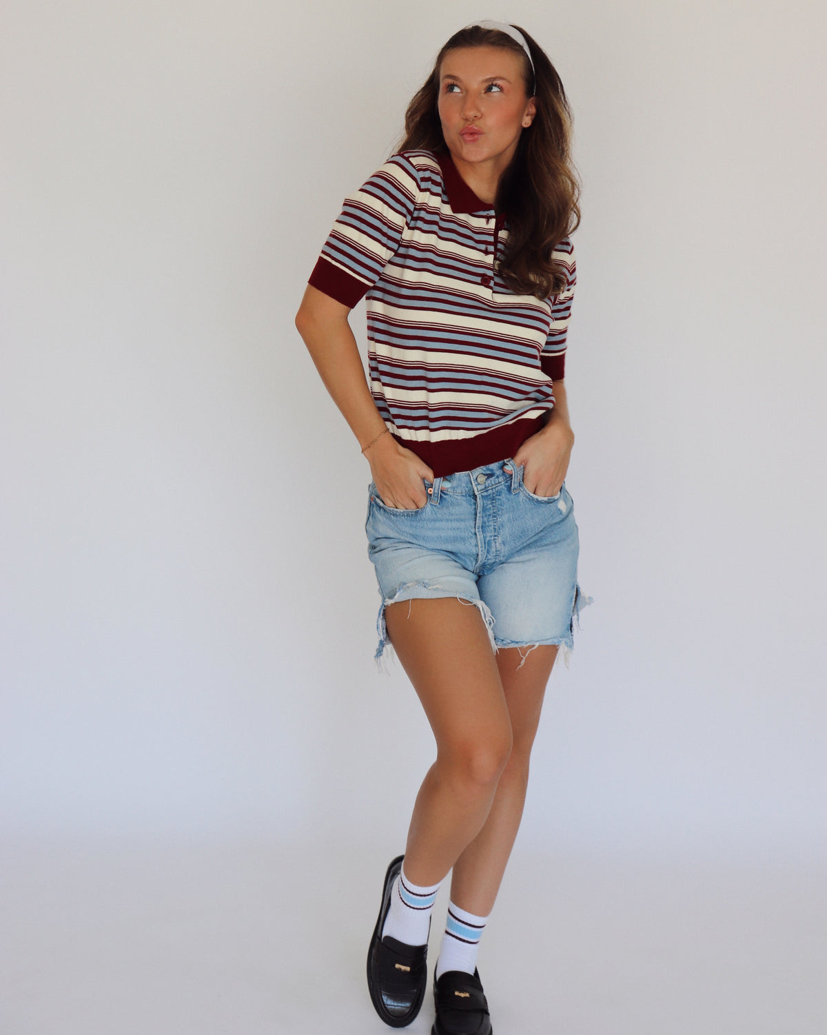 A woman modeling a short-sleeve knit polo shirt with horizontal stripes in cream, burgundy, and blue, paired with light-wash denim shorts, white socks, and black loafers against a plain white studio background.