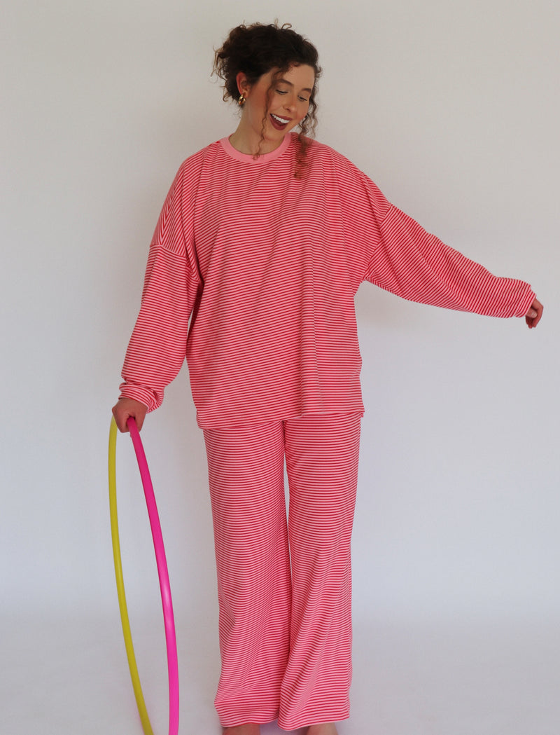 A woman modeling a pink striped lounge set, consisting of a long-sleeve top and matching pants, while holding a pink and yellow and pink hula hoop against a plain white background.