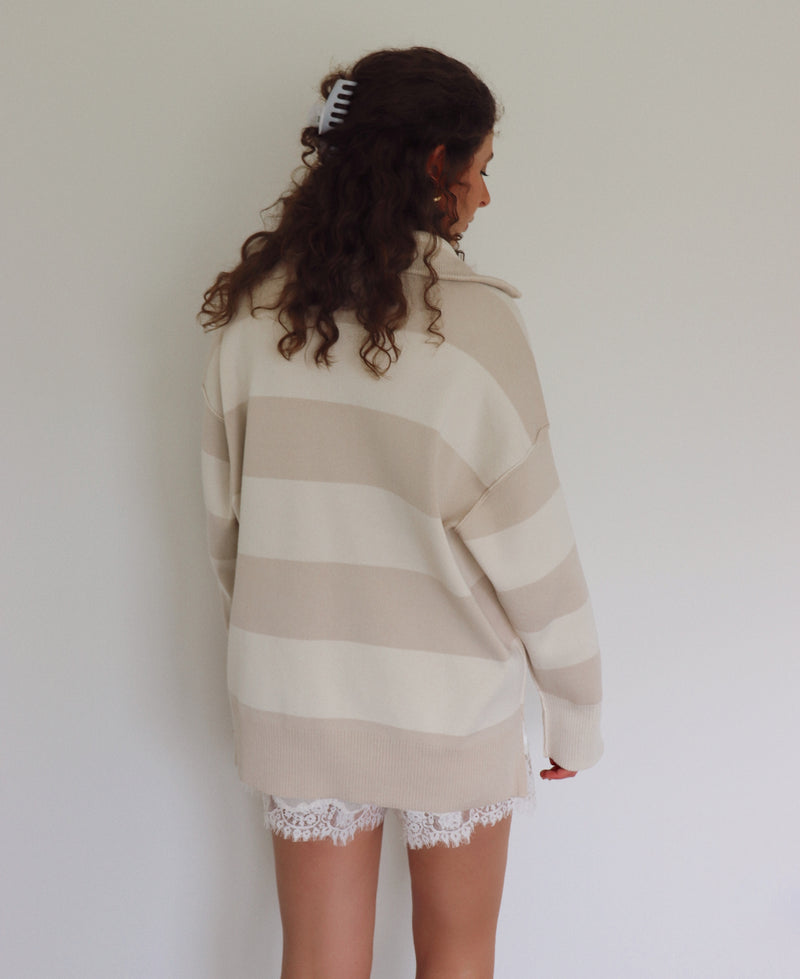 A woman shown from the back wearing an oversized beige and cream striped knit sweater, paired with white lace shorts, against a plain light gray wall.
