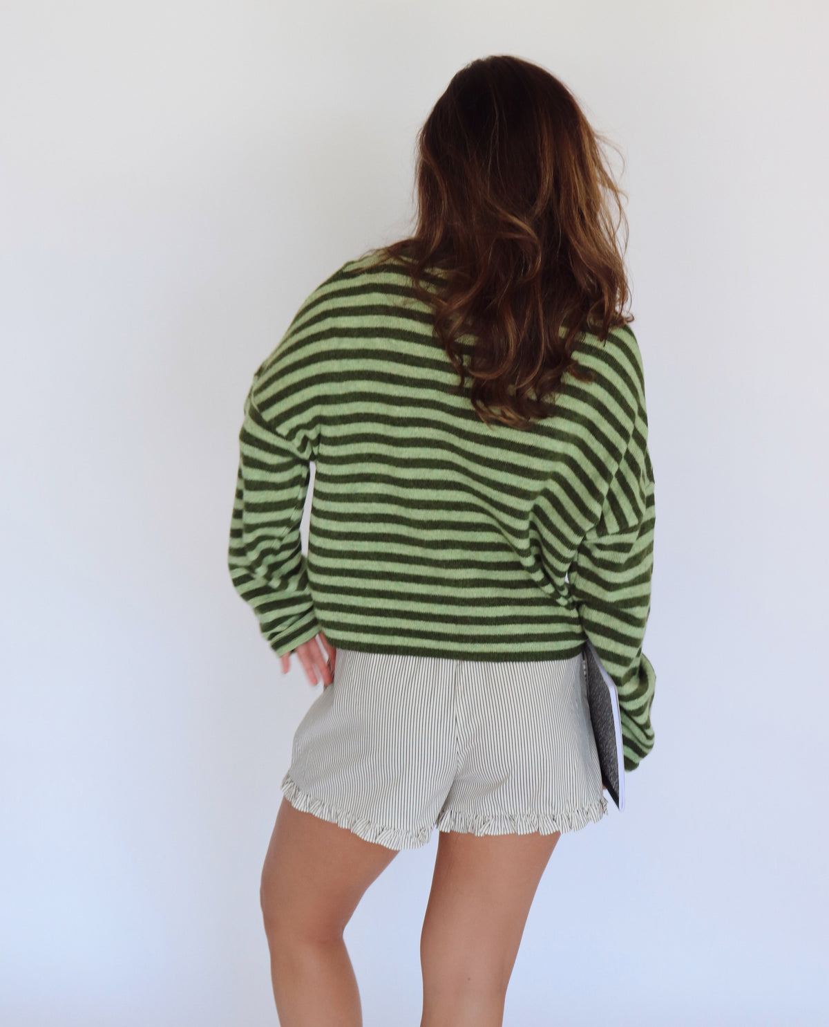 A woman shown from behind wearing a green and dark green striped long-sleeve knit top and light beige shorts, against a plain white studio background.