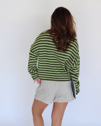 A woman shown from behind wearing a green and dark green striped long-sleeve knit top and light beige shorts, against a plain white studio background.