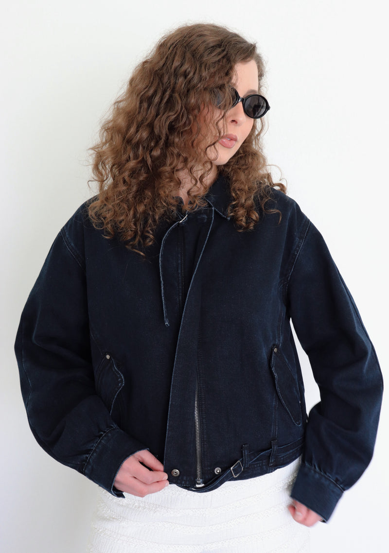 A woman with curly brown hair and sunglasses wearing a dark navy blue cropped denim jacket and white textured shorts, posing against a plain white wall.