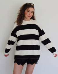A woman with curly brown hair wearing an oversized black and off-white striped long-sleeve sweater and black lace-trim shorts, standing against a plain light gray wall.