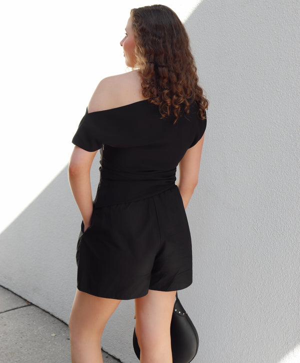 Woman wearing a black off-shoulder top and shorts against a light gray wall.