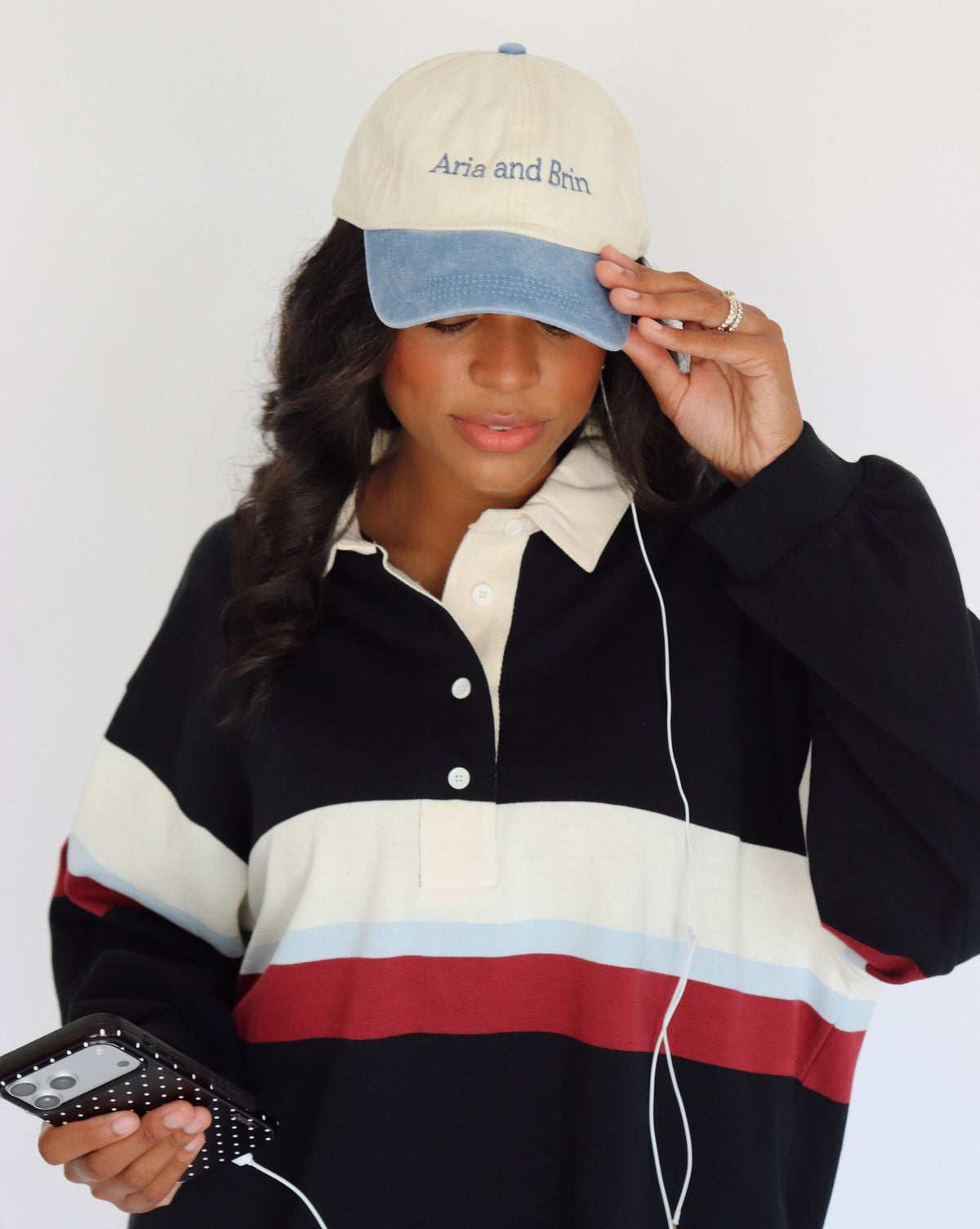 A woman wearing a cream and blue baseball cap with 'Aria and Brin' embroidered on it, paired with a black, cream, light blue, and red color-blocked long-sleeve polo shirt. She is holding a phone with a black polka-dot case and white wired earphones against a plain white background.