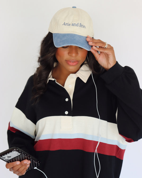 A woman wearing a cream and blue baseball cap with 'Aria and Brin' embroidered on it, paired with a black, cream, light blue, and red color-blocked long-sleeve polo shirt. She is holding a phone with a black polka-dot case and white wired earphones against a plain white background.