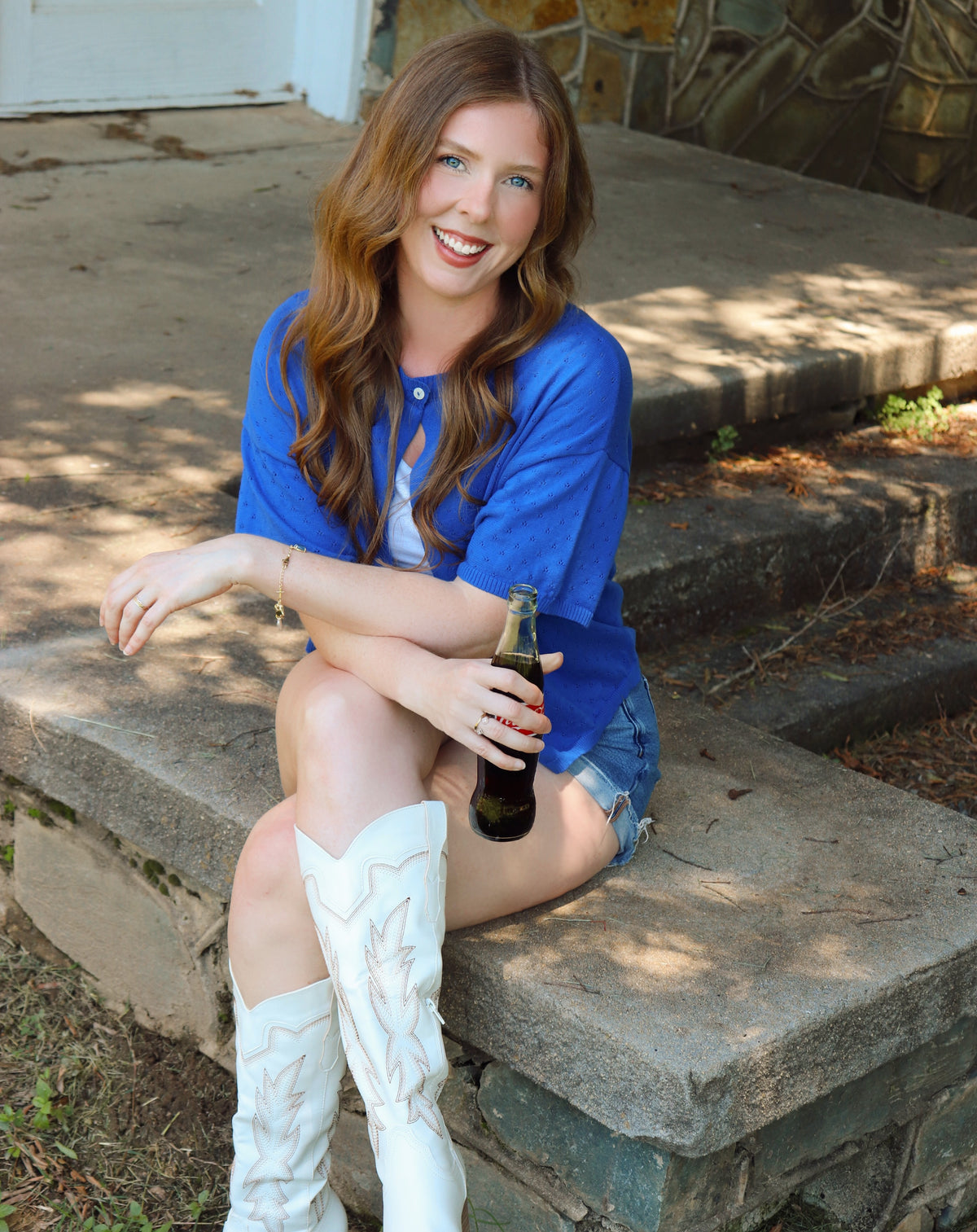 Woman in blue cardigan and white boots sitting on steps holding a bottle.