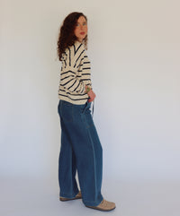 A woman with curly brown hair wearing a cream-colored knit cardigan with navy horizontal stripes and blue jeans, standing against a plain light gray background.