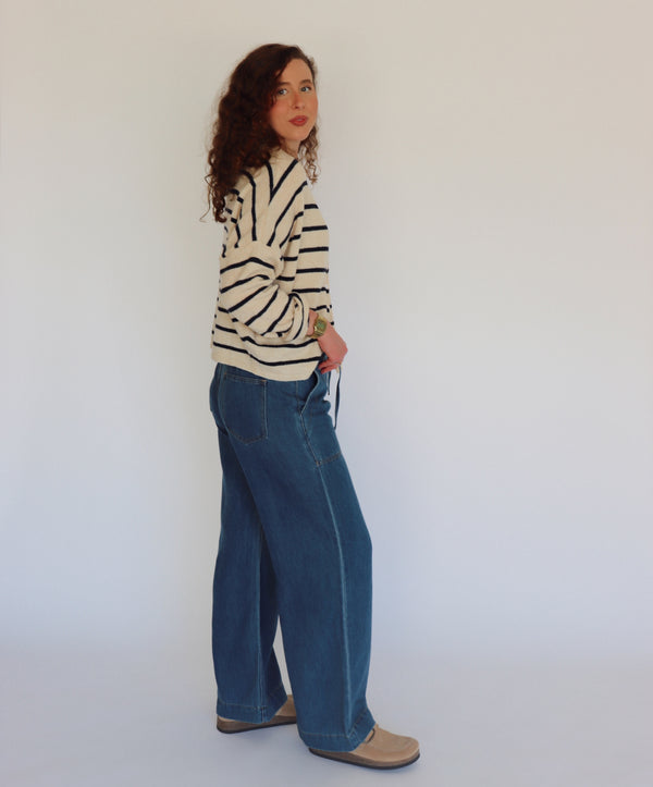A woman with curly brown hair wearing a cream-colored knit cardigan with navy horizontal stripes and blue jeans, standing against a plain light gray background.